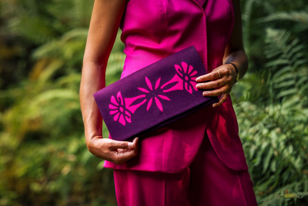 Handgemachte Clutch in Violett mit floralem Muster, gehalten von Frau im pinkfarbenen Outfit – Design von Atelier Kordula Magiera.